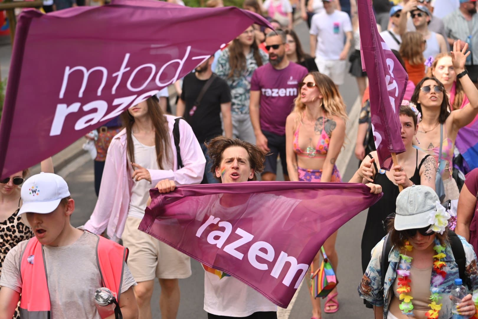 Młody mężczyzna bierze udział w marszu równości. Biegnie, trzymając w dłoniach flagę razem. Ma na policzku namalowaną tęczę. Ma rozwiane włosy. Jest otoczony ludźmi w letnich strojach.