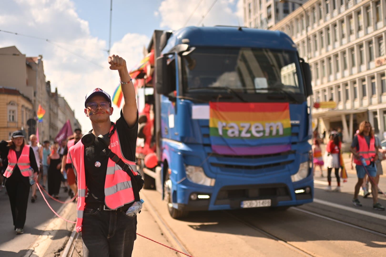Młody chłopak z namalowaną na policzkach tęczką podnosi pięść w górę. Ma na sobie odblaskową kamizelkę. Idzie przed samochodem, który ma na masce tęczową flagę z napisem „razem”.