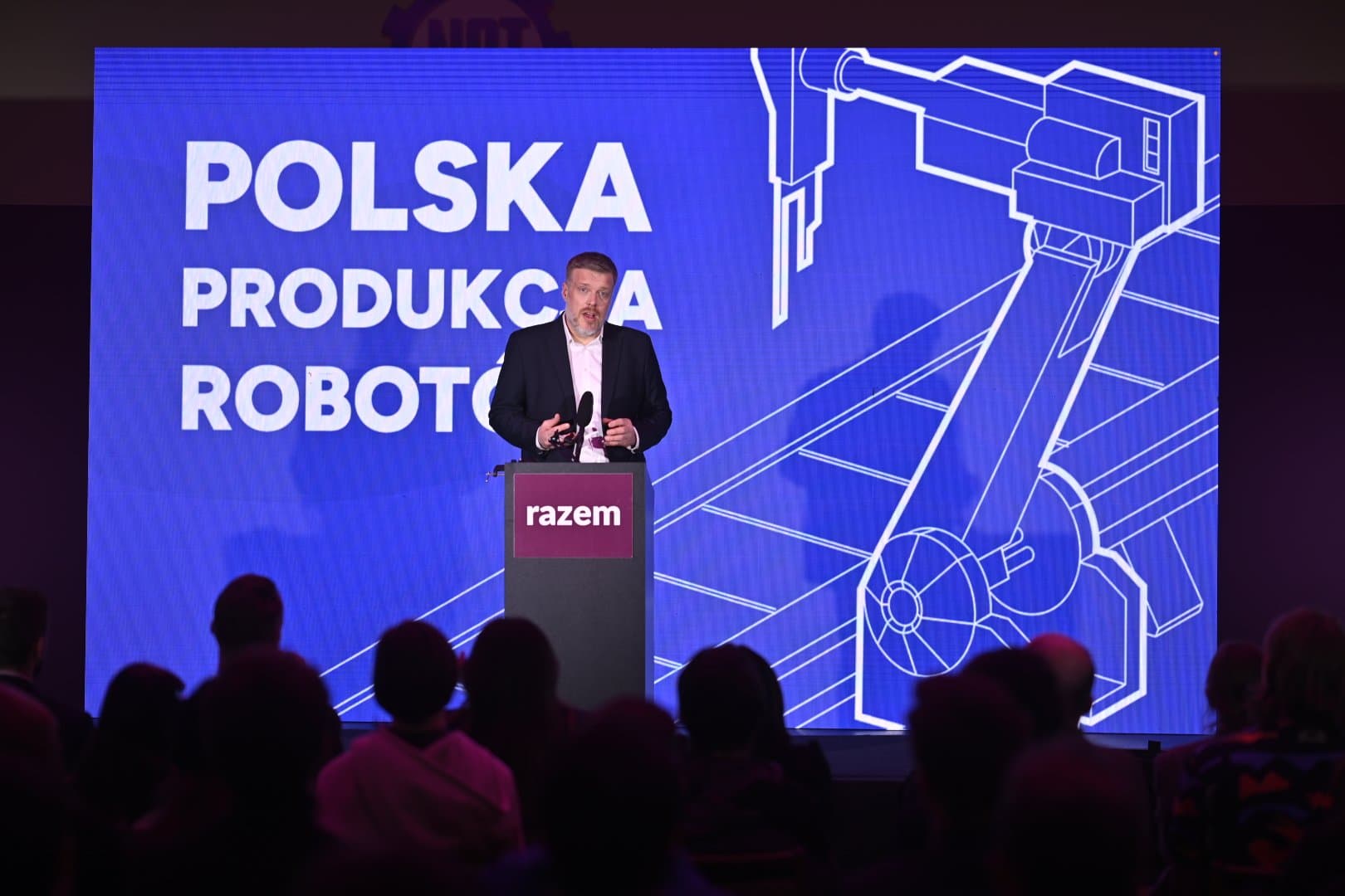 Mówca stoi przy mównicy z logo Razem przed publicznością. Za nim niebieski ekran z napisem „Polska produkcja robotów” i rysunkiem ramienia robota.