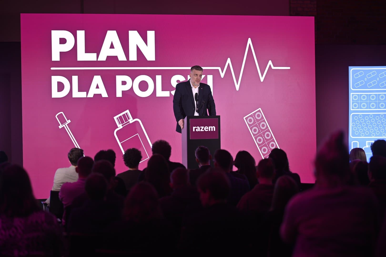 Mówca stoi przy mównicy z logo Razem przed publicznością. Za nim różowy ekran z napisem „Plan dla Polski” i rysunkami leków.