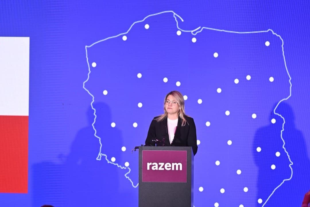Kobieta stoi przy mównicy z logo Razem przed publicznością. Za nią niebieski ekran z konturem mapy Polski i biało-czerwona flaga.