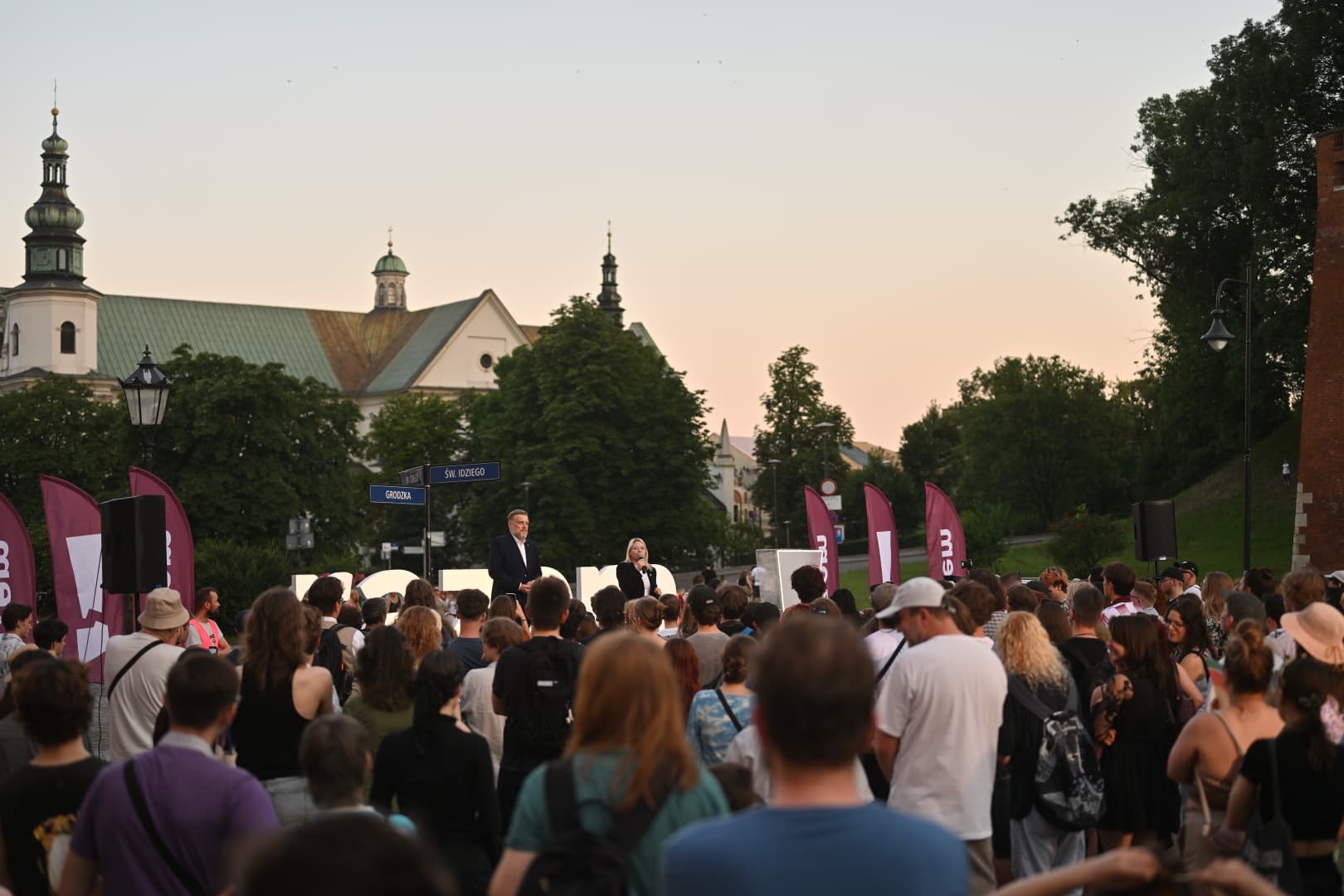 Wiec Razem o zmierzchu w Krakowie, u zbiegu ulic Grodzkiej i św. Idziego. Tłum słucha dwojga mówców na scenie, obok stoją flagi Razem. W tle kościół z zielonymi hełmami wież.